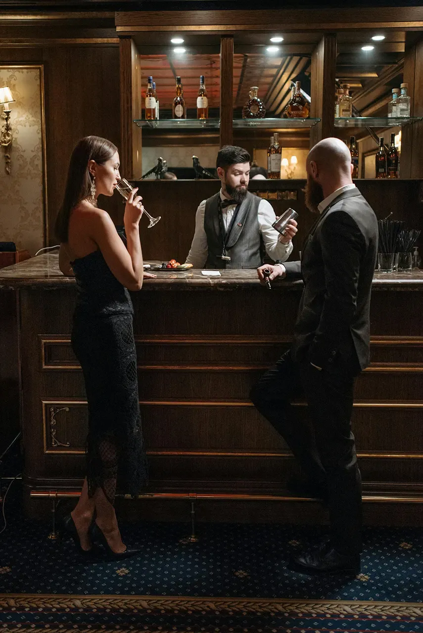 Elegant bar scene with a woman in a black dress sipping champagne, a bartender in a vest preparing a drink, and a man in a suit engaging in conversation, showcasing a sophisticated cocktail experience in a Las Vegas restaurant.